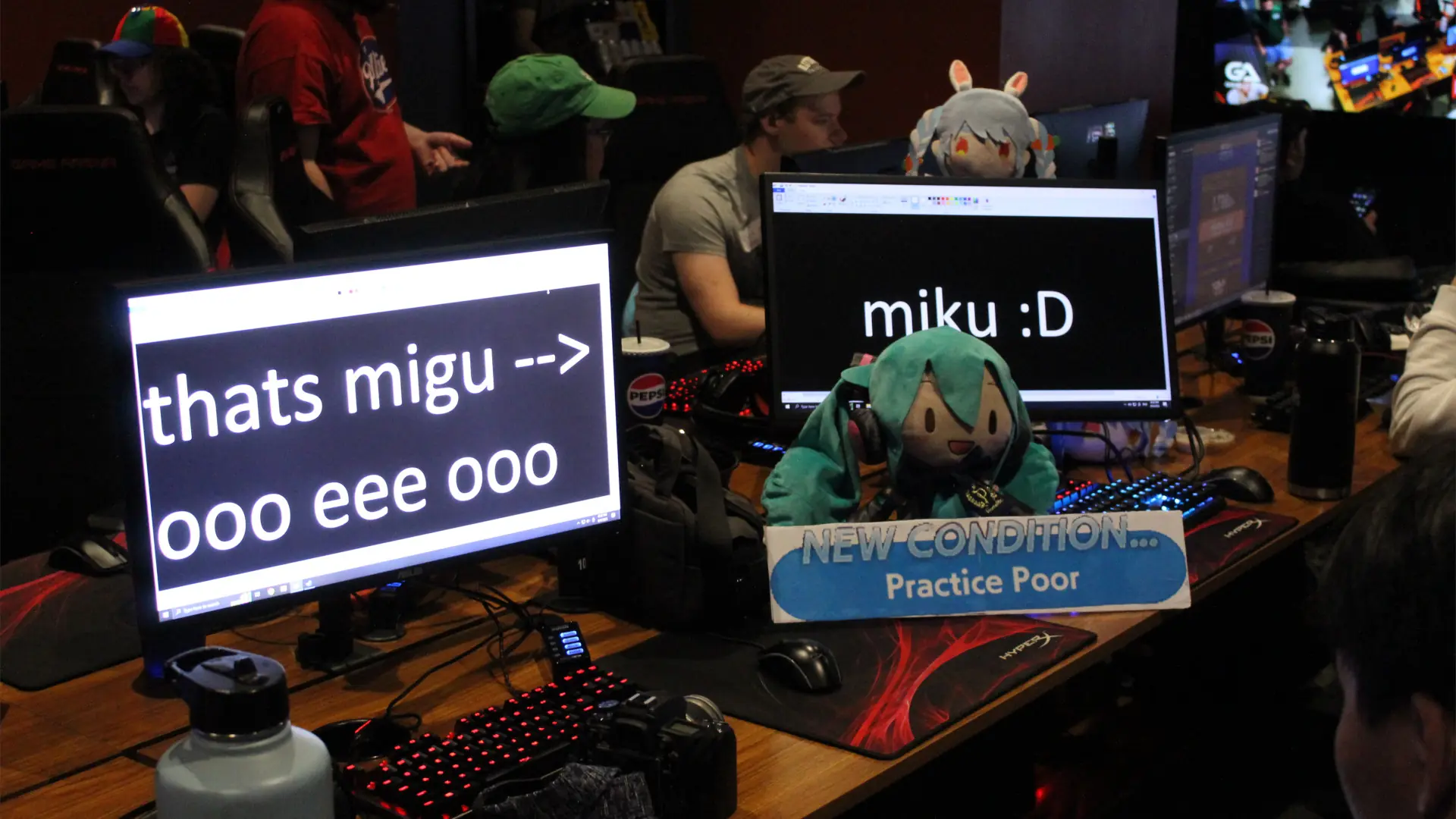 A plush of Hatsune Miku next to two computer monitors with meme text and a 'Practice Poor' sign.